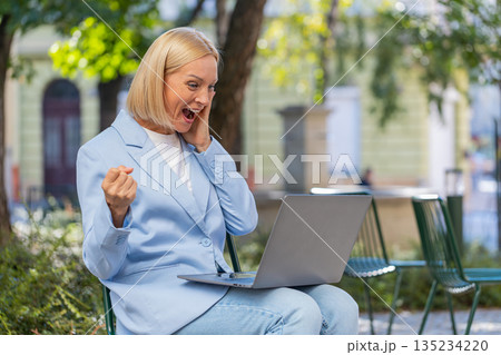 Happy mature businesswoman using laptop computer celebrating success victory clenching fists in city 135234220