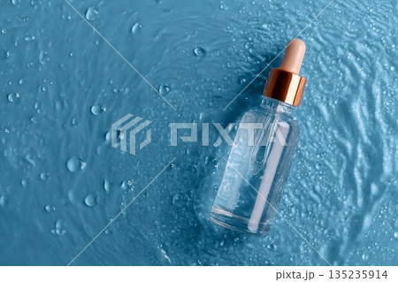 Glass bottle of anti aging serum and falling water drops on blue background. Facial serum mockup. 135235914