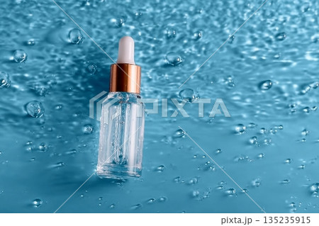Glass bottle of anti aging serum and falling water drops on blue background. Facial serum mockup. 135235915