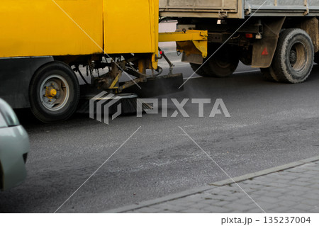 A yellow road sweeper machine is cleaning the asphalt on a city street. The circular brush is visible as it sweeps dust and debris A yellow road sweeper machine is cleaning the asphalt on a city street. The circular brush is visible as it sweeps dust and debris 135237004