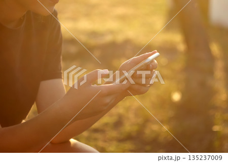 A close-up of a person hands holding and using a smartphone outdoors during the golden hour. using smartphone outdoors golden hour light A close-up of a person hands holding and using a smartphone outdoors during the golden hour. using smartphone outdoors golden hour light 135237009