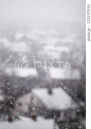 A vertical, blurry shot of snowflakes falling over a snow-covered town. The houses and roofs are out of focus in the background, creating a soft, wintry atmosphere. Vertical snowfall background A vertical, blurry shot of snowflakes falling over a snow-covered town. The houses and roofs are out of focus in the background, creating a soft, wintry atmosphere. Vertical snowfall background 135237010
