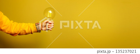 A person arm in a bright yellow sweater holding a glowing LED light bulb against a solid yellow background. Concept of having a great idea, innovation, or creative thinking. Banner with copy space 135237021