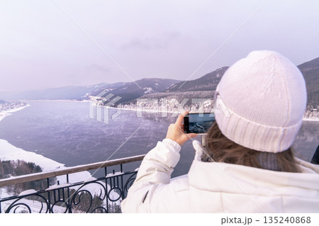 A woman in a white warm jacket takes pictures with her smartphone of a beautiful view of the Yenisei River in Siberia, Russia from the Tsar Fish viewing point near the village, rear view. High quality A woman in a white warm jacket takes pictures with her smartphone of a beautiful view of the Yenisei River in Siberia, Russia from the Tsar Fish viewing point near the village, rear view. High quality 135240868