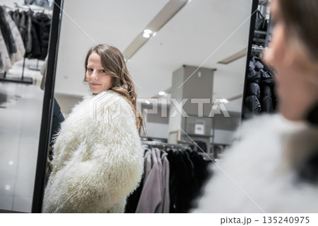 In a clothing boutique, a young woman admires herself in a mirror while trying on a white sheepskin coat. She enjoys seasonal fashion shopping during the holiday time In a clothing boutique, a young woman admires herself in a mirror while trying on a white sheepskin coat. She enjoys seasonal fashion shopping during the holiday time 135240975