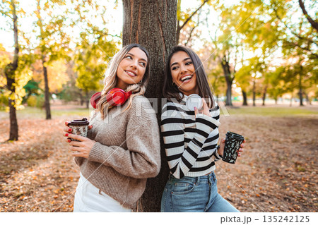 Friends enjoy autumn colors and warm drinks in the park while laughing together Friends enjoy autumn colors and warm drinks in the park while laughing together 135242125