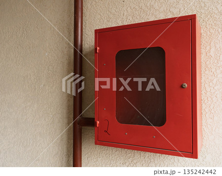 Red metal fire safety cabinet mounted on a textured beige wall. Industrial utility box with pipe and window. Emergency equipment housing Red metal fire safety cabinet mounted on a textured beige wall. Industrial utility box with pipe and window. Emergency equipment housing 135242442
