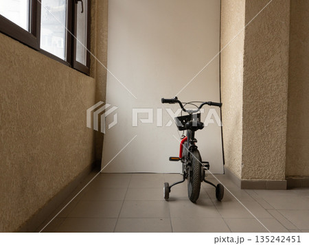Rear view of a black child's bicycle with training wheels indoors. Small bike with basket and stabilizers on a balcony. Copy space on white background Rear view of a black child's bicycle with training wheels indoors. Small bike with basket and stabilizers on a balcony. Copy space on white background 135242451