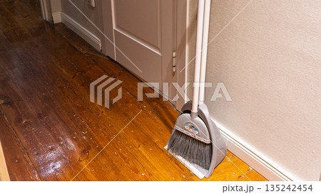 Grey broom and dustpan set standing on worn wooden floor. Domestic cleaning tools against beige wall in hallway. Housework and chores concept Grey broom and dustpan set standing on worn wooden floor. Domestic cleaning tools against beige wall in hallway. Housework and chores concept 135242454