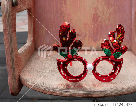 Novelty Christmas glasses with red sequined reindeer antlers on a rustic wooden seat. Festive holiday party accessory and costume prop 135242478