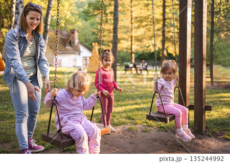 Mother is rocking her twin daughters on a swing in the park Mother is rocking her twin daughters on a swing in the park 135244992