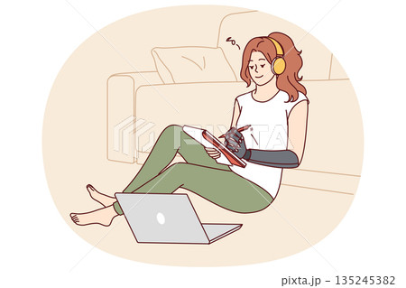 Woman with bionic prosthetic arm sits on floor near sofa and makes notes in notebook Woman with bionic prosthetic arm sits on floor near sofa and makes notes in notebook 135245382