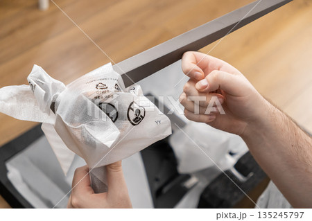 Hands carefully unboxing a new sleek silver monitor stand from protective plastic film, displaying recycling symbols and OTHER waste instructions 135245797