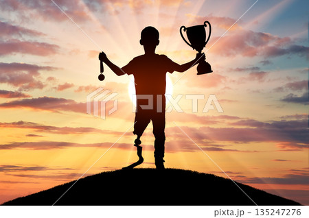 A young boy with a prosthetic leg stands victoriously on a hilltop, holding a trophy and a medal 135247276
