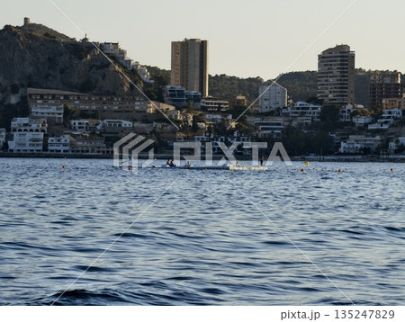 lone boat moves peacefully across quiet bay with city lights in distance during evening twilight 135247829