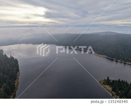 Calm Forest Lake At Sunrise With Haze, Quiet Wilderness Scene Featuring Reflections And Distant Hills 135248340