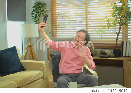 Senior man smiling and waving during a video call on smartphone at home, enjoying digital communication Senior man smiling and waving during a video call on smartphone at home, enjoying digital communication 135249011