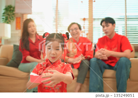 Young girl opens a red envelope gift while her family chats happily together on background Young girl opens a red envelope gift while her family chats happily together on background 135249051