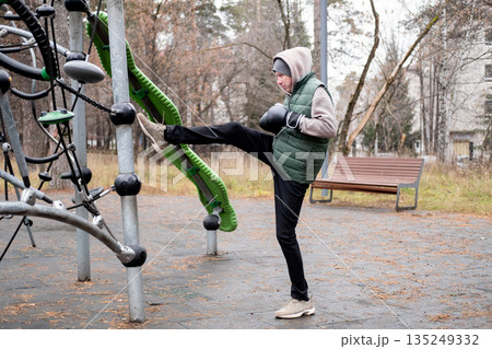 A man in a tracksuit and hat stretches with boxing gloves on an outdoor sports field. Exercising in cold weather. A man in a tracksuit and hat stretches with boxing gloves on an outdoor sports field. Exercising in cold weather. 135249332