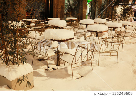 Empty outdoor cafe seating covered with fresh snow during winter evening. The image conveys pause in city life, hospitality concept, and seasonal atmosphere Empty outdoor cafe seating covered with fresh snow during winter evening. The image conveys pause in city life, hospitality concept, and seasonal atmosphere 135249609