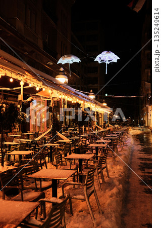 Outdoor cafe terrace covered with snow glows under warm lights at night. The image reflects winter city life, hospitality atmosphere, and seasonal urban charm Outdoor cafe terrace covered with snow glows under warm lights at night. The image reflects winter city life, hospitality atmosphere, and seasonal urban charm 135249614