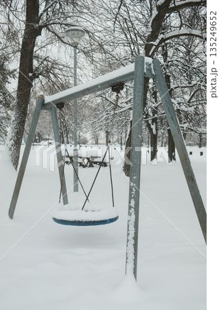 Snow covered playground swing stands empty in a winter park. The scene reflects childhood memories, cold season silence, and urban outdoor life during snowfall. 135249652