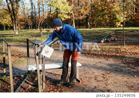 A man adjusts weights on an old outdoor gym machine surrounded by autumn leaves. The concept explores motivation and everyday commitment to physical improvement 135249656