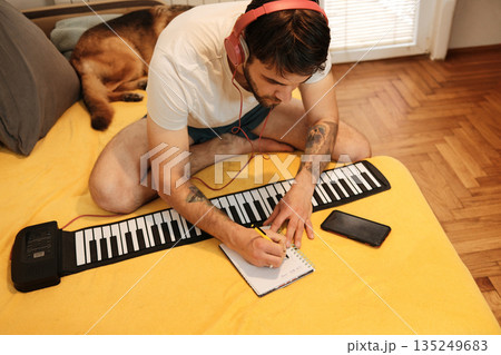 A man writes in a notebook while sitting on a bed with a roll-up keyboard and headphones. The scene reflects structured creative work and the concept of home music development A man writes in a notebook while sitting on a bed with a roll-up keyboard and headphones. The scene reflects structured creative work and the concept of home music development 135249683
