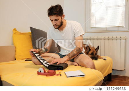 A man sits on a bed and practices on a small roll-up keyboard while a dog rests nearby. The scene shows a quiet creative moment and the concept of home music work A man sits on a bed and practices on a small roll-up keyboard while a dog rests nearby. The scene shows a quiet creative moment and the concept of home music work 135249688