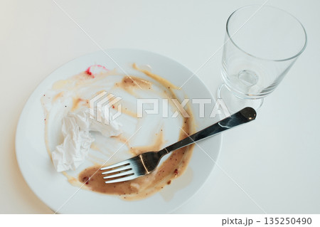 White plate with sauce smears, a crumpled napkin, and an empty glass after a finished meal on a bright table White plate with sauce smears, a crumpled napkin, and an empty glass after a finished meal on a bright table 135250490