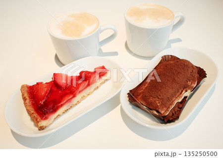 Two desserts, strawberry tart and tiramisu, paired with cups of cappuccino on a white minimalist table under warm light. The concept of breakfast in a cafe Two desserts, strawberry tart and tiramisu, paired with cups of cappuccino on a white minimalist table under warm light. The concept of breakfast in a cafe 135250500