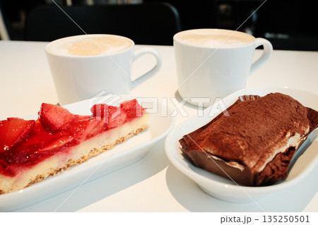 Two desserts, strawberry tart and tiramisu, paired with cups of cappuccino on a white minimalist table under warm light. The concept of breakfast in a cafe Two desserts, strawberry tart and tiramisu, paired with cups of cappuccino on a white minimalist table under warm light. The concept of breakfast in a cafe 135250501