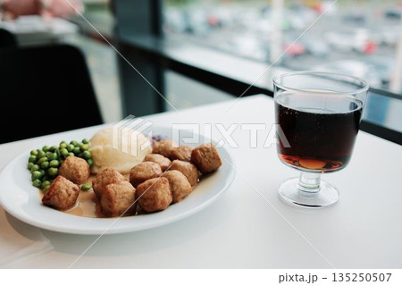 Plate of Swedish meatballs with mashed potatoes, green peas, and gravy served beside a glass of soda on a white table by the window 135250507