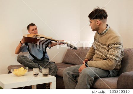 One man shows his guitar to a friend while sitting on a couch at home. The concept reflects creativity, music sharing, and friendly communication One man shows his guitar to a friend while sitting on a couch at home. The concept reflects creativity, music sharing, and friendly communication 135250519