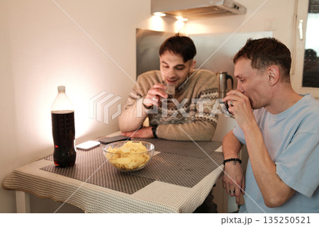 Two friends sit at a kitchen table with snacks and drinks, talking casually. The concept focuses on connection, simple social time, and authentic everyday living Two friends sit at a kitchen table with snacks and drinks, talking casually. The concept focuses on connection, simple social time, and authentic everyday living 135250521