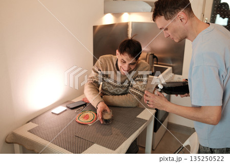 A young man pours a dark soda drink into a glass while his friend smiles at the kitchen table. The concept highlights friendship, casual home moments, and relaxed social interaction A young man pours a dark soda drink into a glass while his friend smiles at the kitchen table. The concept highlights friendship, casual home moments, and relaxed social interaction 135250522