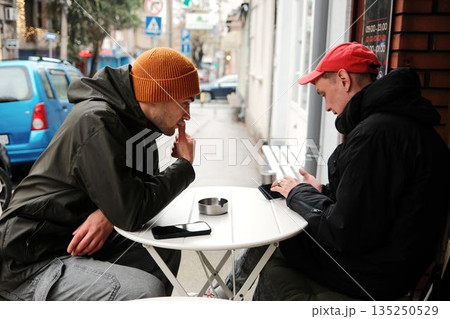 Two men sit at a small outdoor table on a rainy street while focusing on their phones. The scene conveys modern communication concept and urban lifestyle concept Two men sit at a small outdoor table on a rainy street while focusing on their phones. The scene conveys modern communication concept and urban lifestyle concept 135250529
