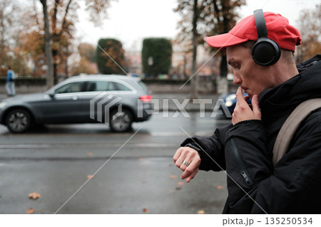 A man in a red cap smokes and checks the time while standing near a busy street on a rainy autumn day. The scene illustrates the concept of time management and daily planning 135250534