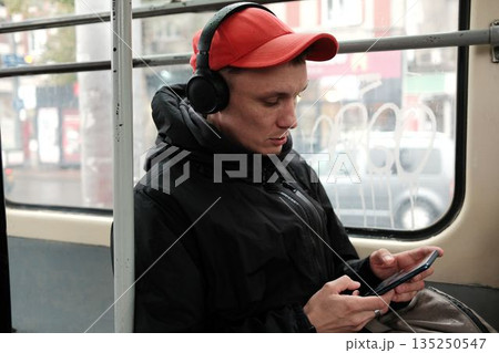 A man sits in a tram wearing headphones and uses his phone. The scene expresses urban commute and digital connection concept A man sits in a tram wearing headphones and uses his phone. The scene expresses urban commute and digital connection concept 135250547