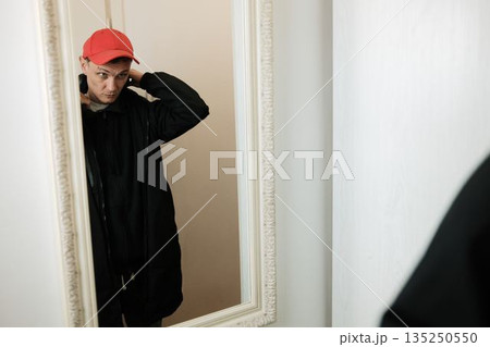 A man adjusts a red cap on his head while looking into the mirror. The moment reflects personal care and self presentation concept A man adjusts a red cap on his head while looking into the mirror. The moment reflects personal care and self presentation concept 135250550