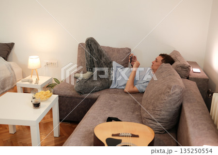 A young man lies on a sofa using his smartphone in a relaxed position. The moment illustrates the concept of leisure and digital habits A young man lies on a sofa using his smartphone in a relaxed position. The moment illustrates the concept of leisure and digital habits 135250554