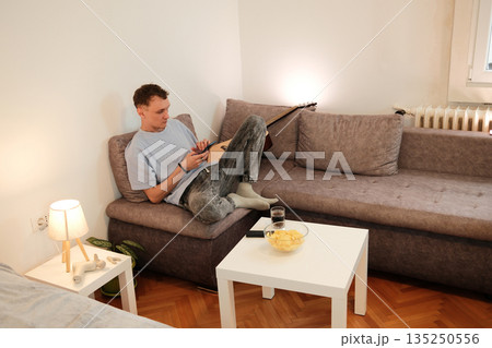 A man lounges on a sofa while typing on his smartphone with a relaxed posture. The scene expresses the concept of digital communication and personal downtime 135250556