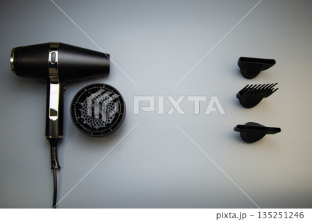 A black hair dryer and a diffuser nozzle lie on one side, on the other there are drying attachments - a comb and classic nozzles of different diameters on a gray background, a top view with space for A black hair dryer and a diffuser nozzle lie on one side, on the other there are drying attachments - a comb and classic nozzles of different diameters on a gray background, a top view with space for 135251246