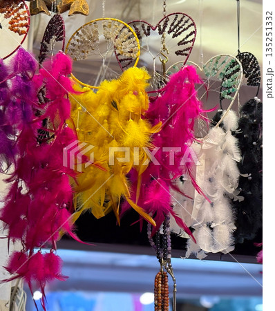 Bright dreamcatchers with colorful feathers hanging in rows at a market stall. Culture, decoration and symbolic spirituality expressed through handmade craft, tradition and consumer creativity. Bright dreamcatchers with colorful feathers hanging in rows at a market stall. Culture, decoration and symbolic spirituality expressed through handmade craft, tradition and consumer creativity. 135251332