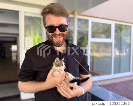 Man in sunglasses smiling and holding small kitten outdoors. Companionship affection, and emotional bond between human and animal. 135251366