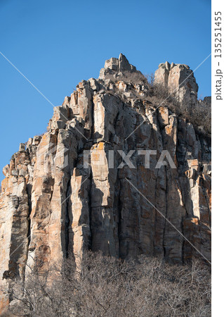 Dramatic rocky cliff rises against a clear blue sky in Armenia, vertical stone formations, warm earth tones, rugged mountain landscape Dramatic rocky cliff rises against a clear blue sky in Armenia, vertical stone formations, warm earth tones, rugged mountain landscape 135251455