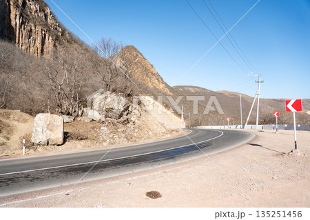 Curved mountain road runs through rocky landscape in Armenia, dry hillsides, clear blue sky, road signs and power lines emphasizing direction, travel Curved mountain road runs through rocky landscape in Armenia, dry hillsides, clear blue sky, road signs and power lines emphasizing direction, travel 135251456