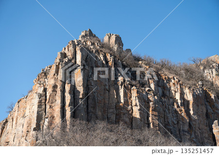 Dramatic rocky cliff rises against a clear blue sky in Armenia, vertical stone formations, warm earth tones, rugged mountain landscape Dramatic rocky cliff rises against a clear blue sky in Armenia, vertical stone formations, warm earth tones, rugged mountain landscape 135251457
