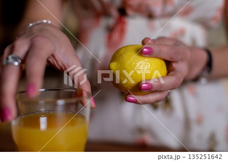 Hand holding lemon above drink, Closeup of lemon and hand preparing juice, Natural light illuminates hand and lemon during everyday juice preparation in cozy kitchen Hand holding lemon above drink, Closeup of lemon and hand preparing juice, Natural light illuminates hand and lemon during everyday juice preparation in cozy kitchen 135251642