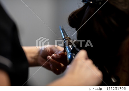 Closeup stylist curling dark hair with iron, hands guiding tool, warm lighting, focused technique, texture and shine emphasized, intimate behindthescenes salon moment 135251706